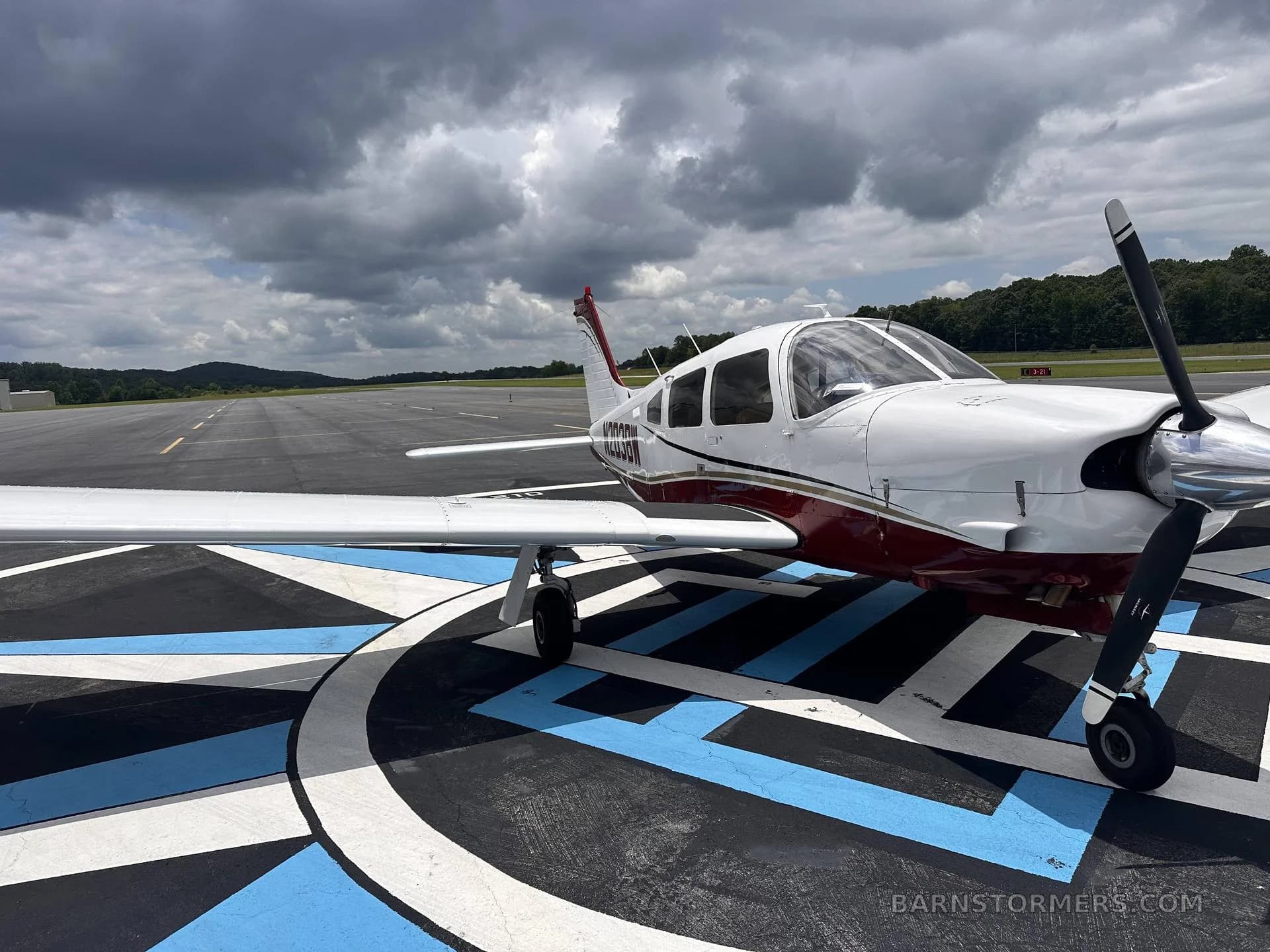 A white and maroon single-engine propeller aircraft (N203BW) is parked on a blue and white patterned tarmac with a runway in the background under a cloudy sky.