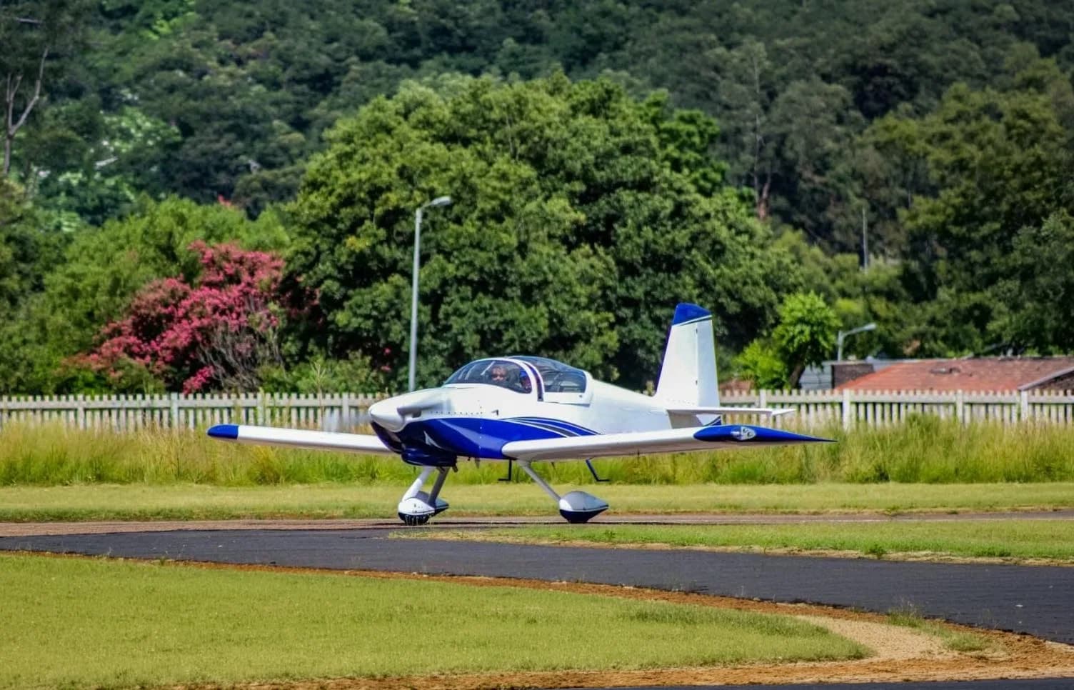 A white and blue single-engine aircraft with a bubble canopy is seen on a tarmac with green trees and a fence in the background.