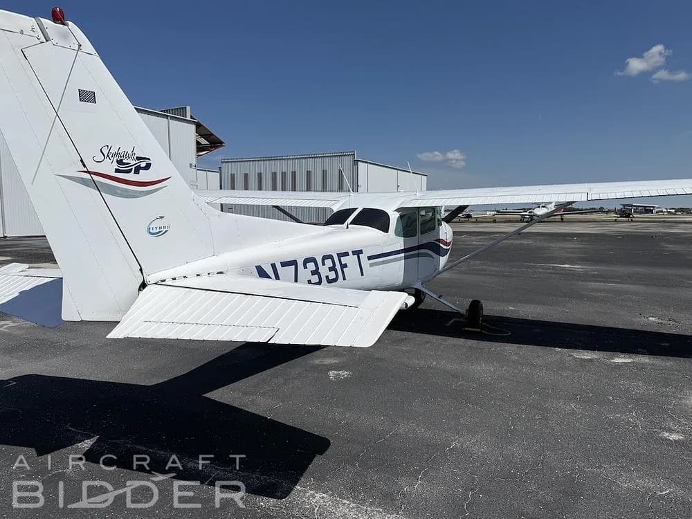 A rear view of the white Cessna Skyhawk N733FT parked near hangars on a sunny day.