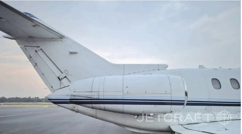 A close-up view of the rear fuselage, engine, and tail of a white business jet with blue stripes, parked on an airfield.
