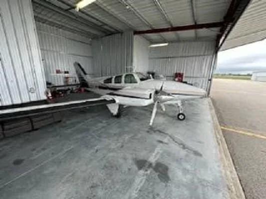 A white and blue twin-engine aircraft, N55EZ, is parked inside a hangar with the door open, viewed from a wider front-right angle.