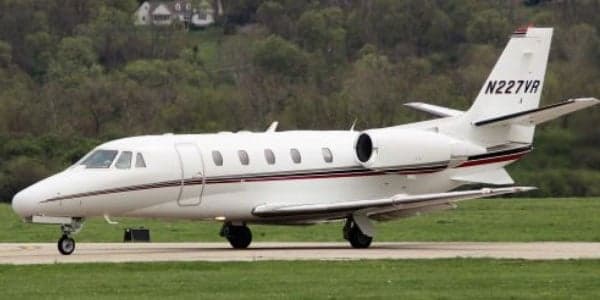 A white Cessna Citation jet with a red and black stripe is parked on the grass.