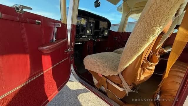 The open door and front right seat of a small aircraft, showing red interior trim and a sheepskin seat cover.