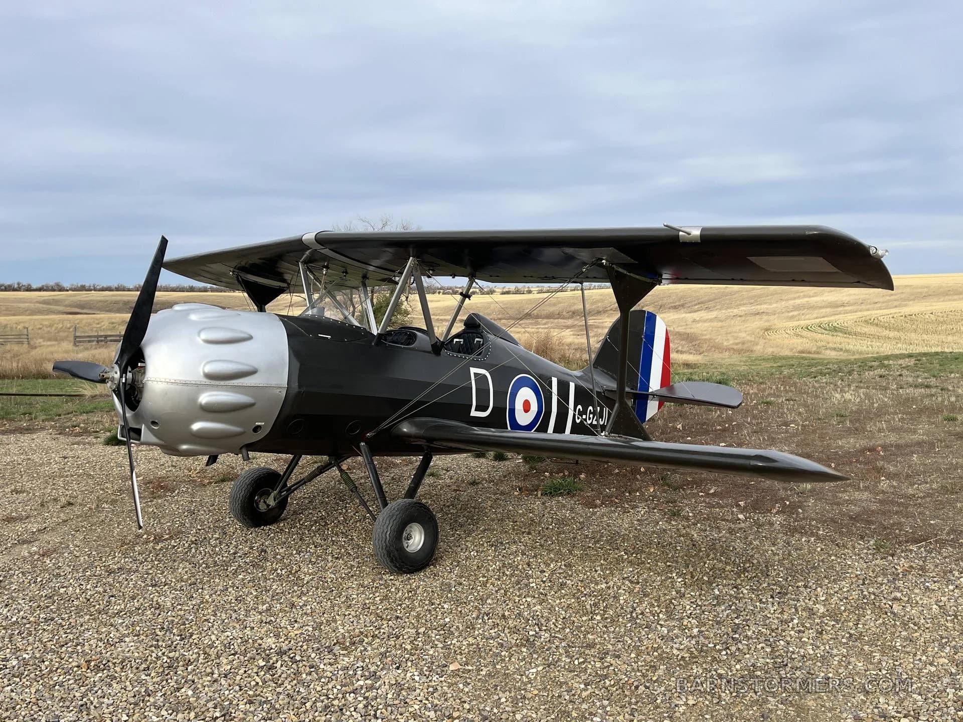 A black biplane with a silver engine cowling and British roundel markings, viewed from the front-right, parked on a gravel path in a field.