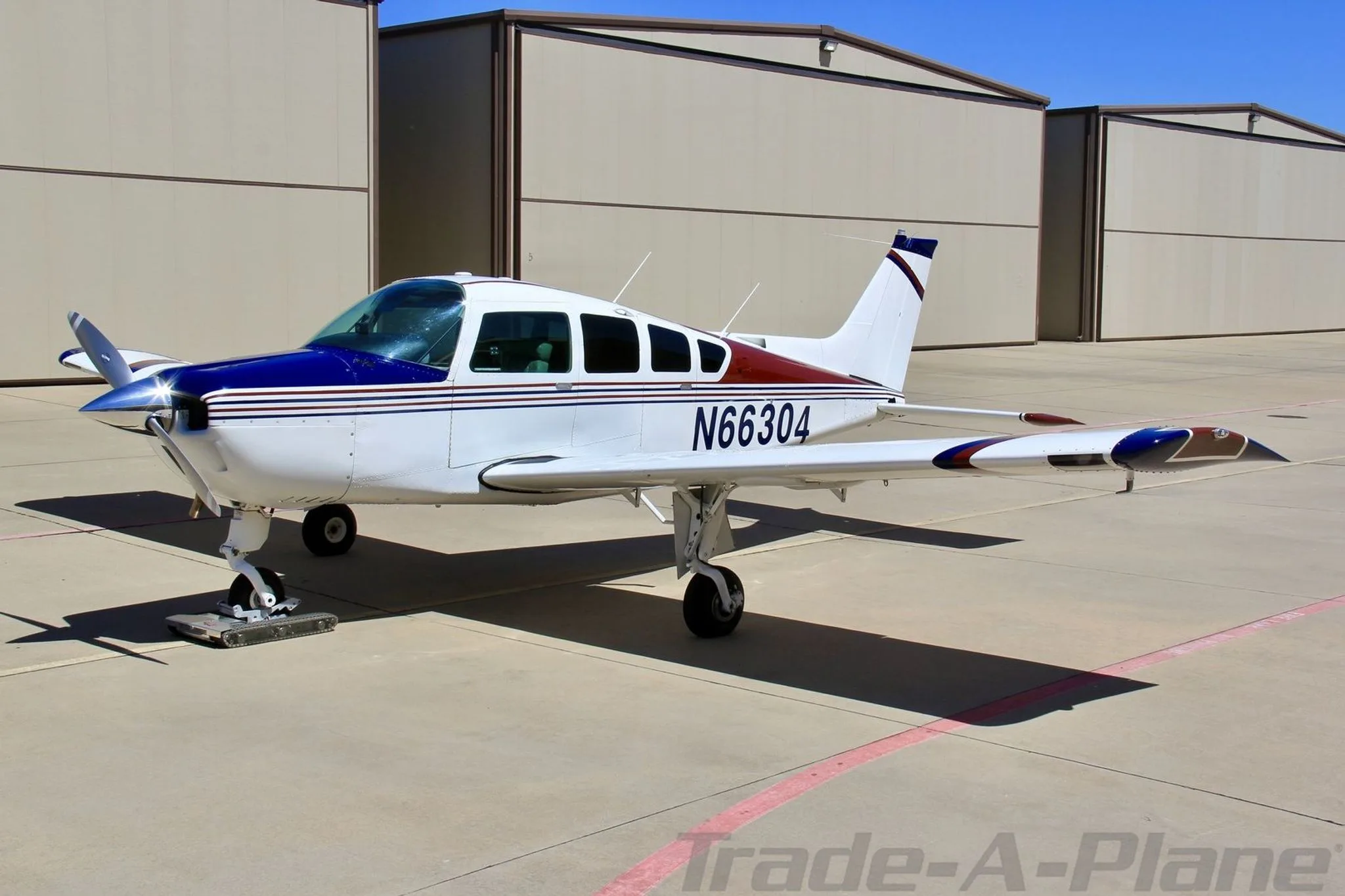 A white, blue, and red single-engine aircraft, N66304, parked on an asphalt apron in front of hangars.