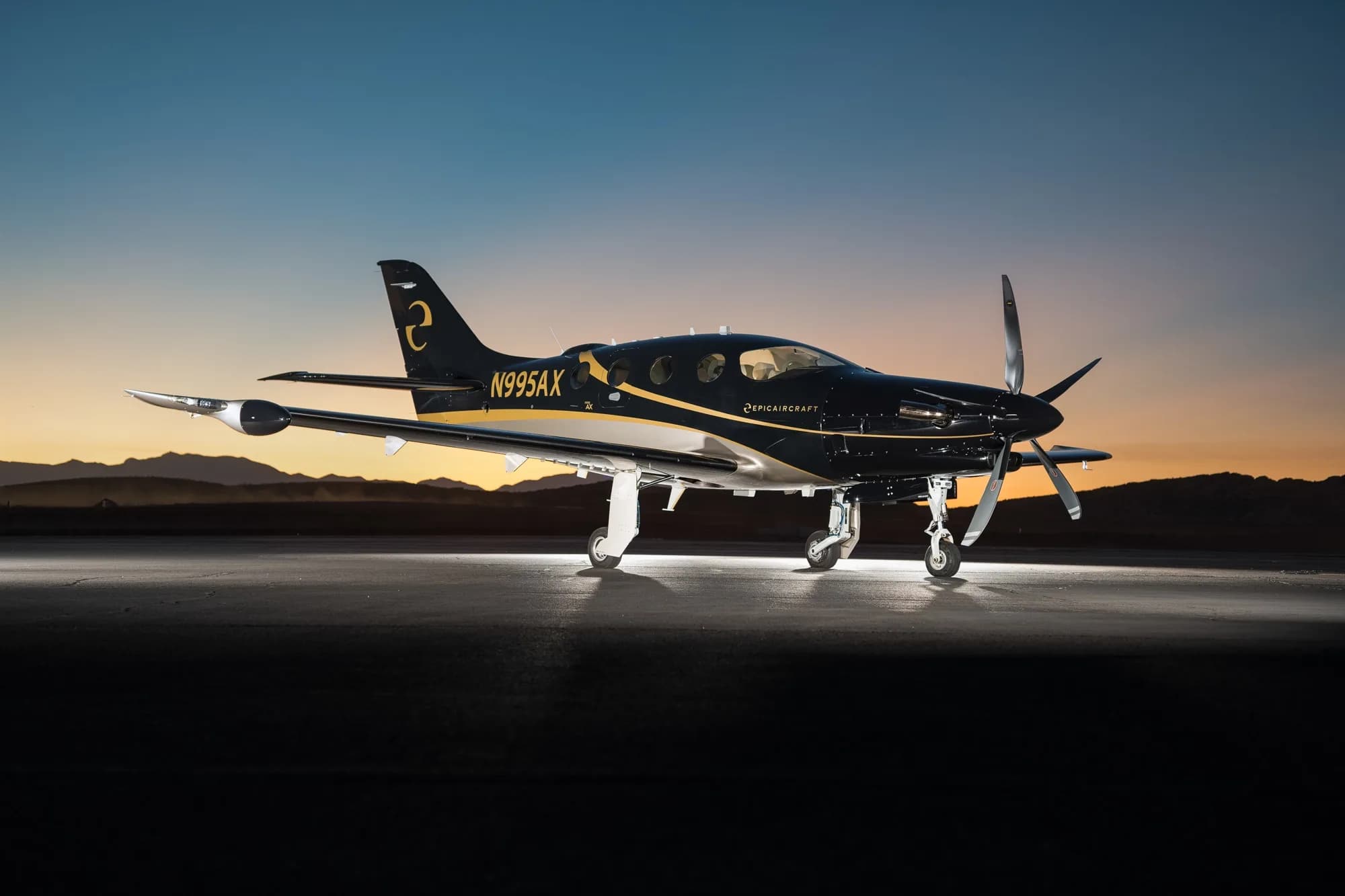A black and gold Epic E1000 GX aircraft (N995AX) is parked on a tarmac at sunset, with mountains in the background.