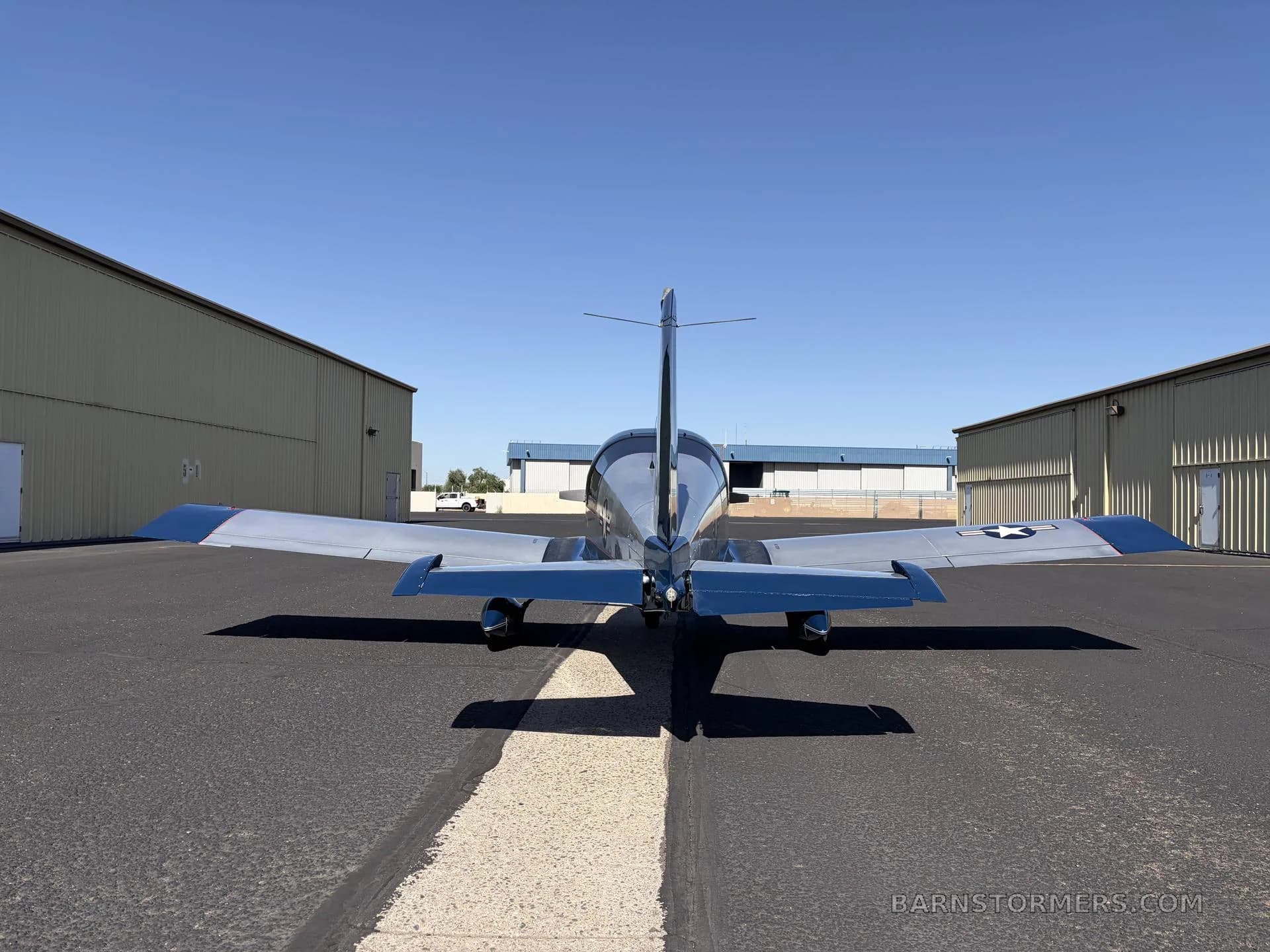 A rear view of a silver and blue Grumman American AA-5 Traveler aircraft with US Air Force star insignia on the wings, parked between hangars.