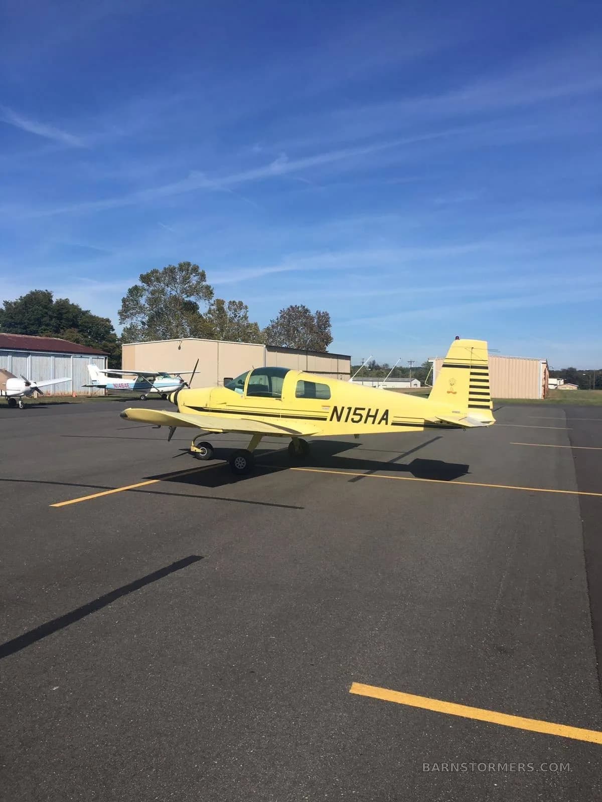 A bright yellow Grumman American AA-1 light aircraft with black stripes and registration N15HA parked on an airport tarmac under a clear blue sky.