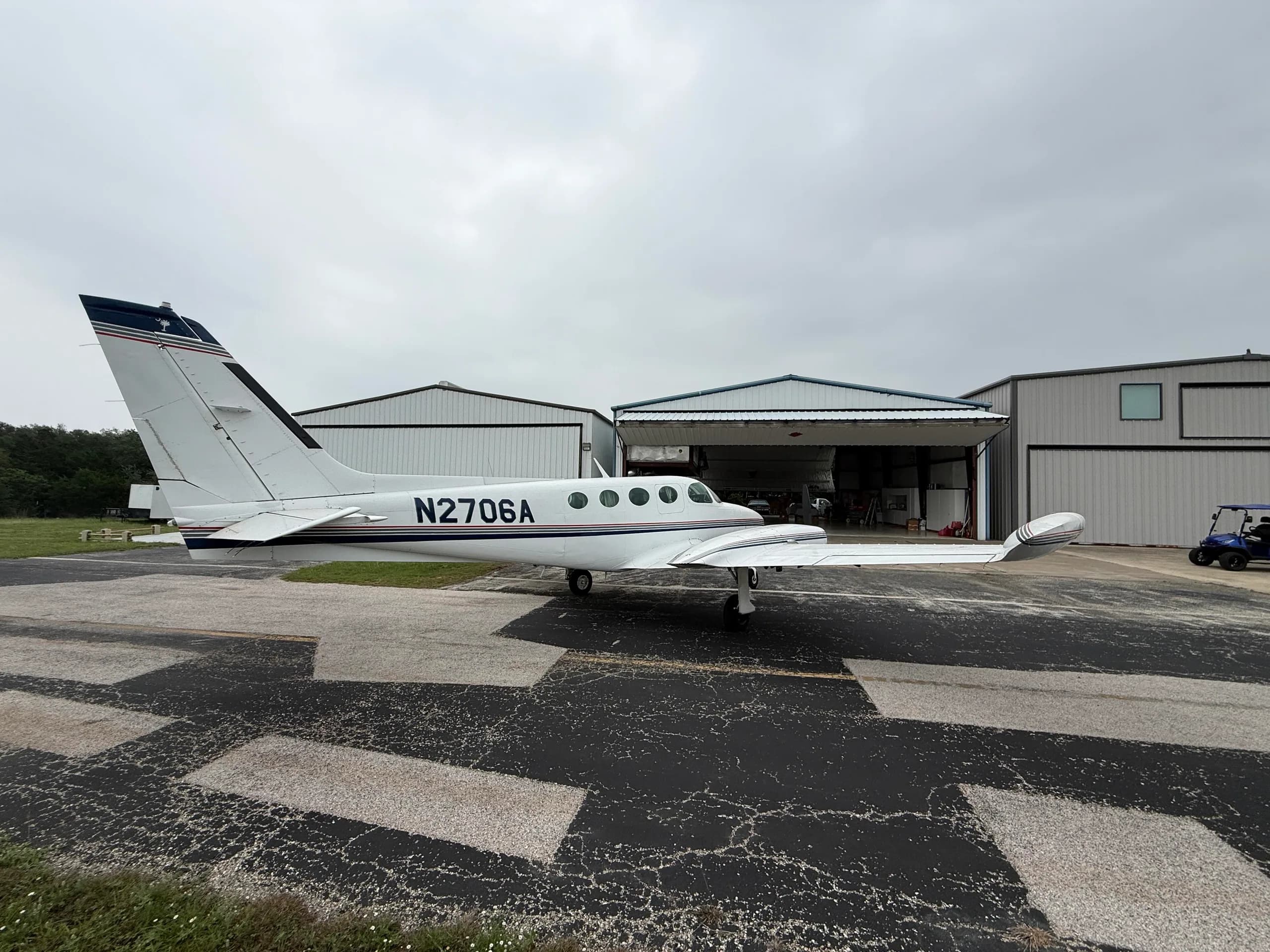 A white Cessna 335 aircraft, registration N2706A, parked on an airfield in front of hangars.
