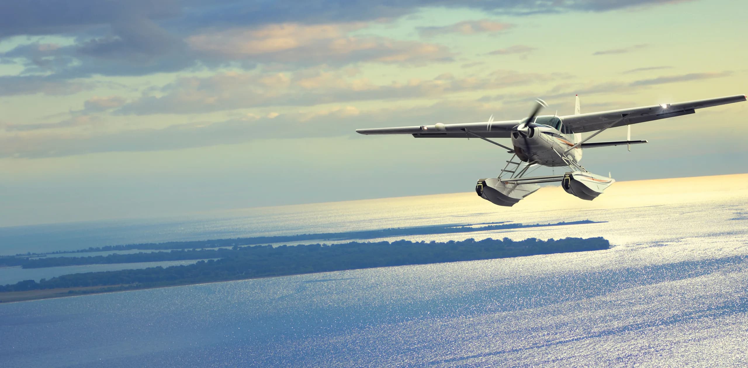 A seaplane flies over the water at sunset.