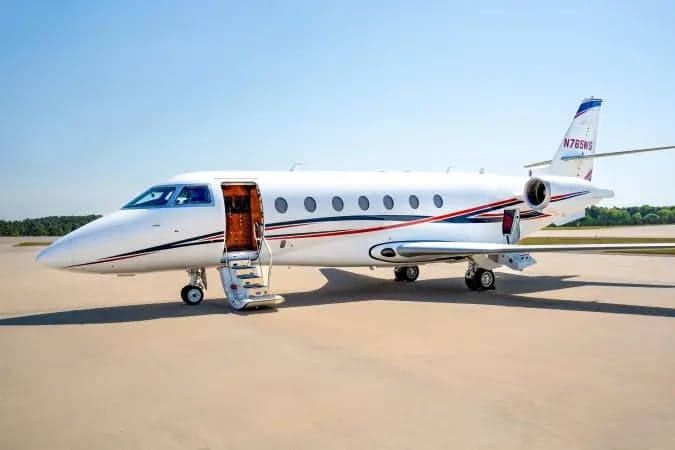 A white business jet with red and blue stripes, registration N765WS, parked on an airfield with its main door open and stairs extended.