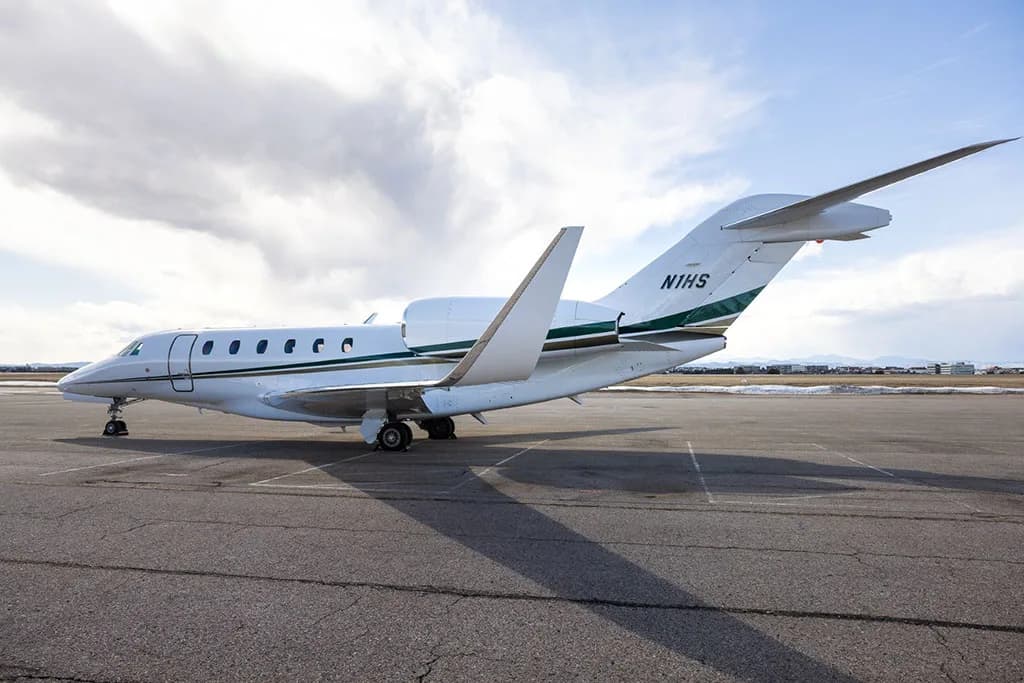 A white business jet with green and gold stripes on the tail, registration N1HS, parked on an airfield under a partly cloudy sky.