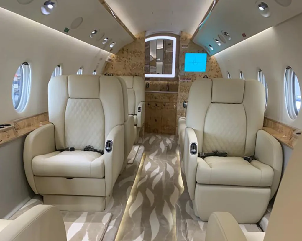 The luxurious interior of a private jet cabin with cream-colored quilted leather seats and a patterned carpet.