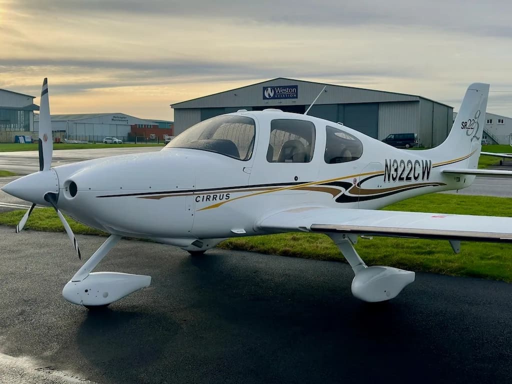 A front-side view of a white Cirrus SR22 aircraft with N322CW registration, parked on a tarmac with hangars under a cloudy sky.