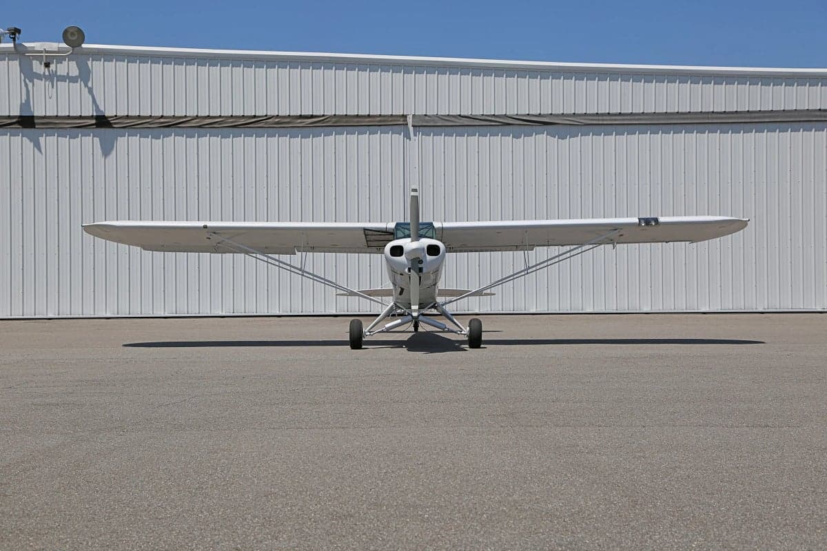 Front view of a white and blue Piper Cub in front of a hangar.