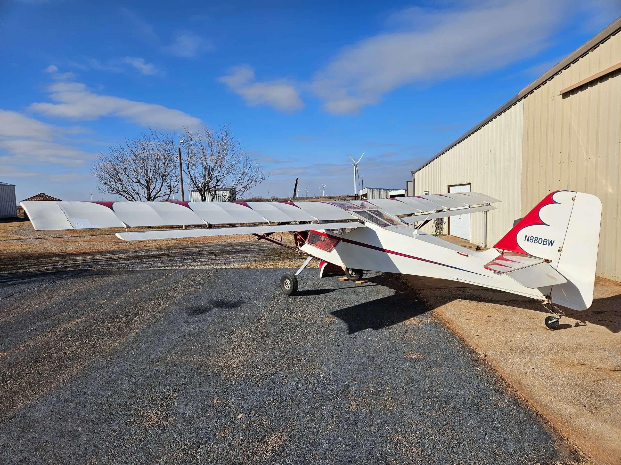 A rear view of the white and red experimental aircraft N880BW parked outside a hangar, with wind turbines visible in the distant background.