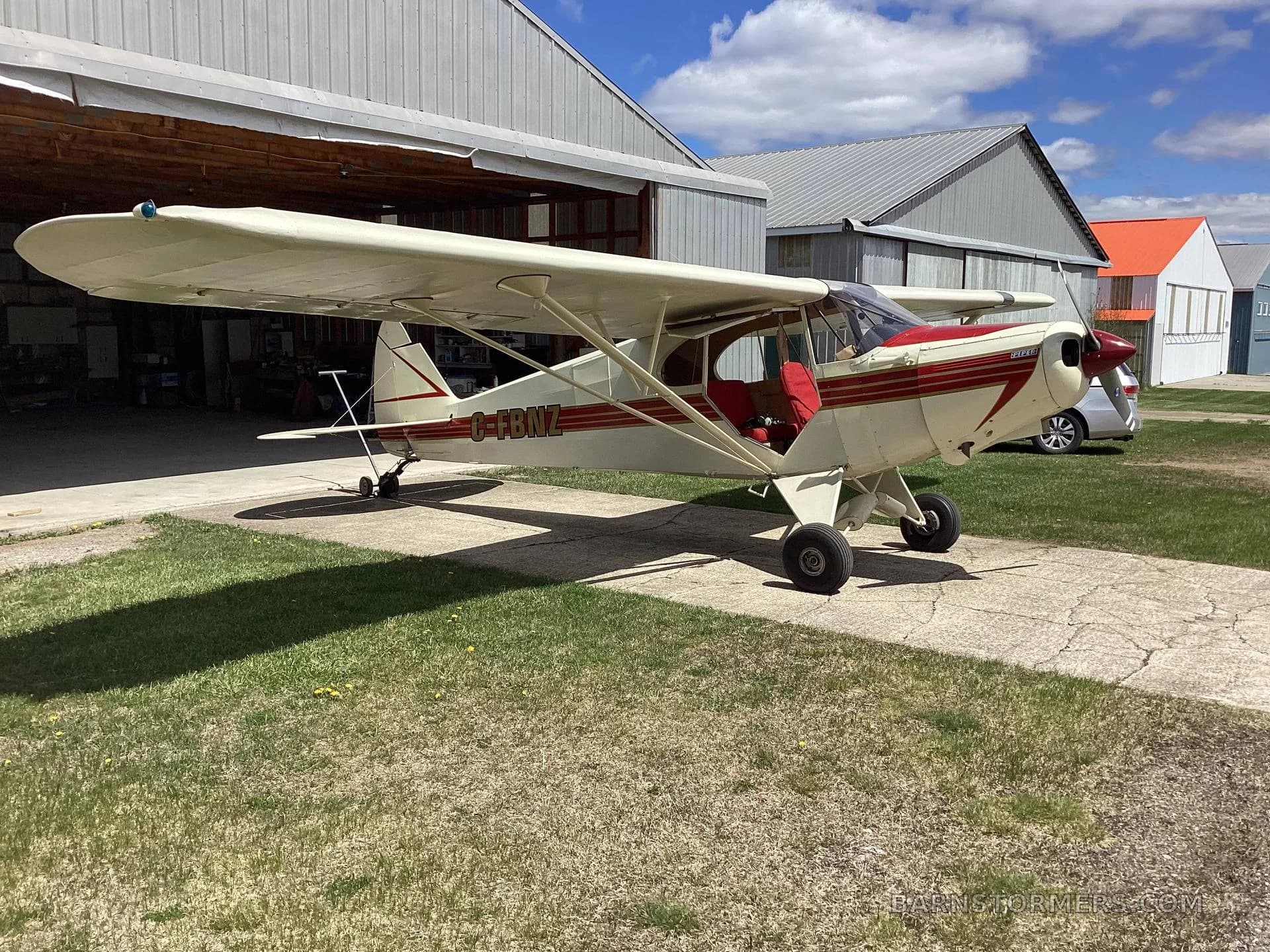 A cream and red high-wing aircraft with tail number C-FBNZ parked on a concrete pad outside a hangar, viewed from the front-right.