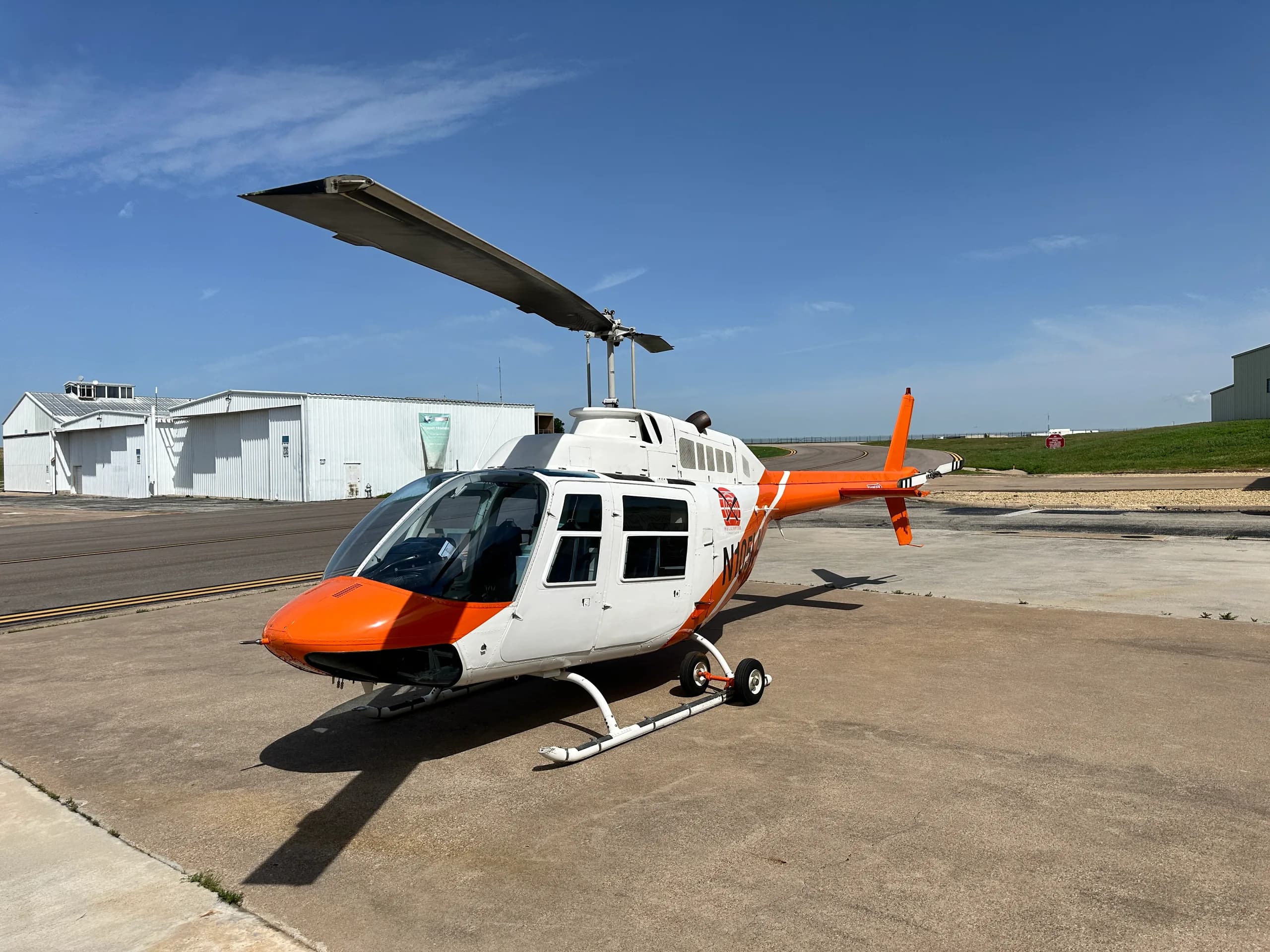 A white and orange helicopter, N1054, parked on an airfield with hangars under a blue sky.