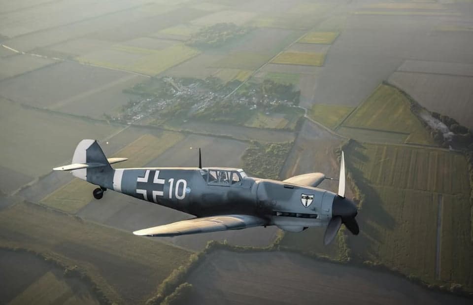 A gray Messerschmitt Bf 109 aircraft in flight, viewed from below.