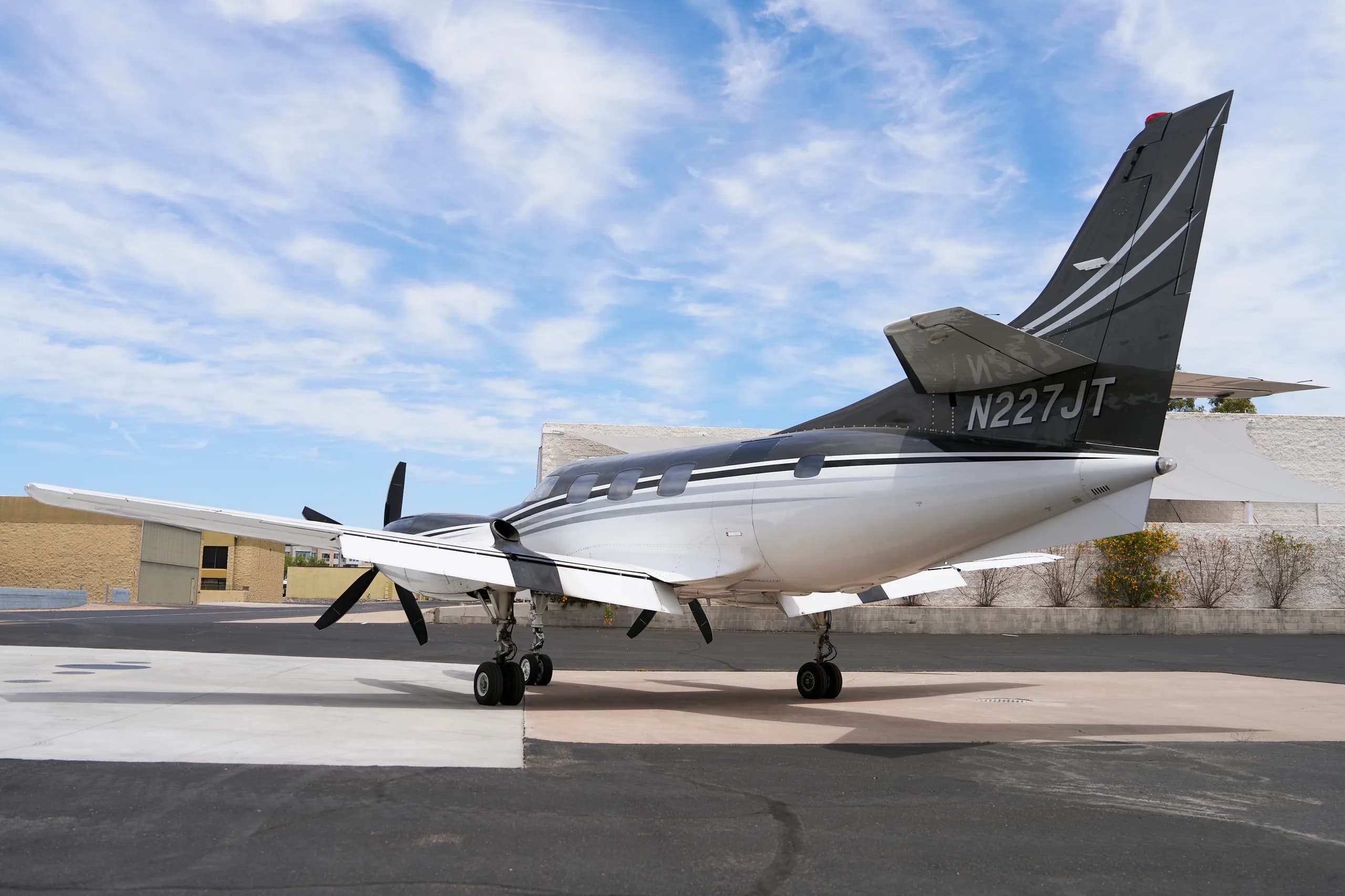 A rear-quarter view of the white and black twin-engine turboprop aircraft, N227JT, parked on a tarmac under a partly cloudy sky.