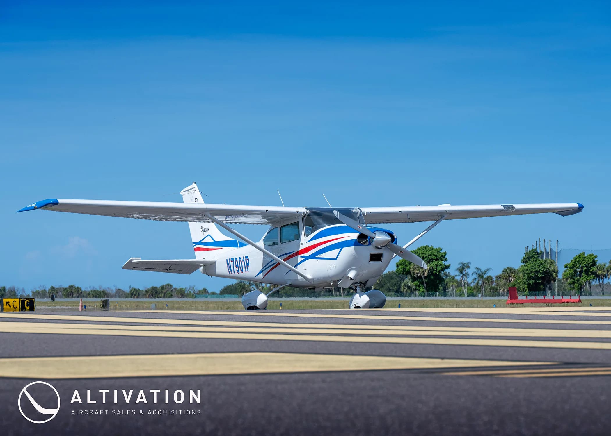 A white Cessna 182 Skylane with blue and red stripes on a runway under a clear blue sky.
