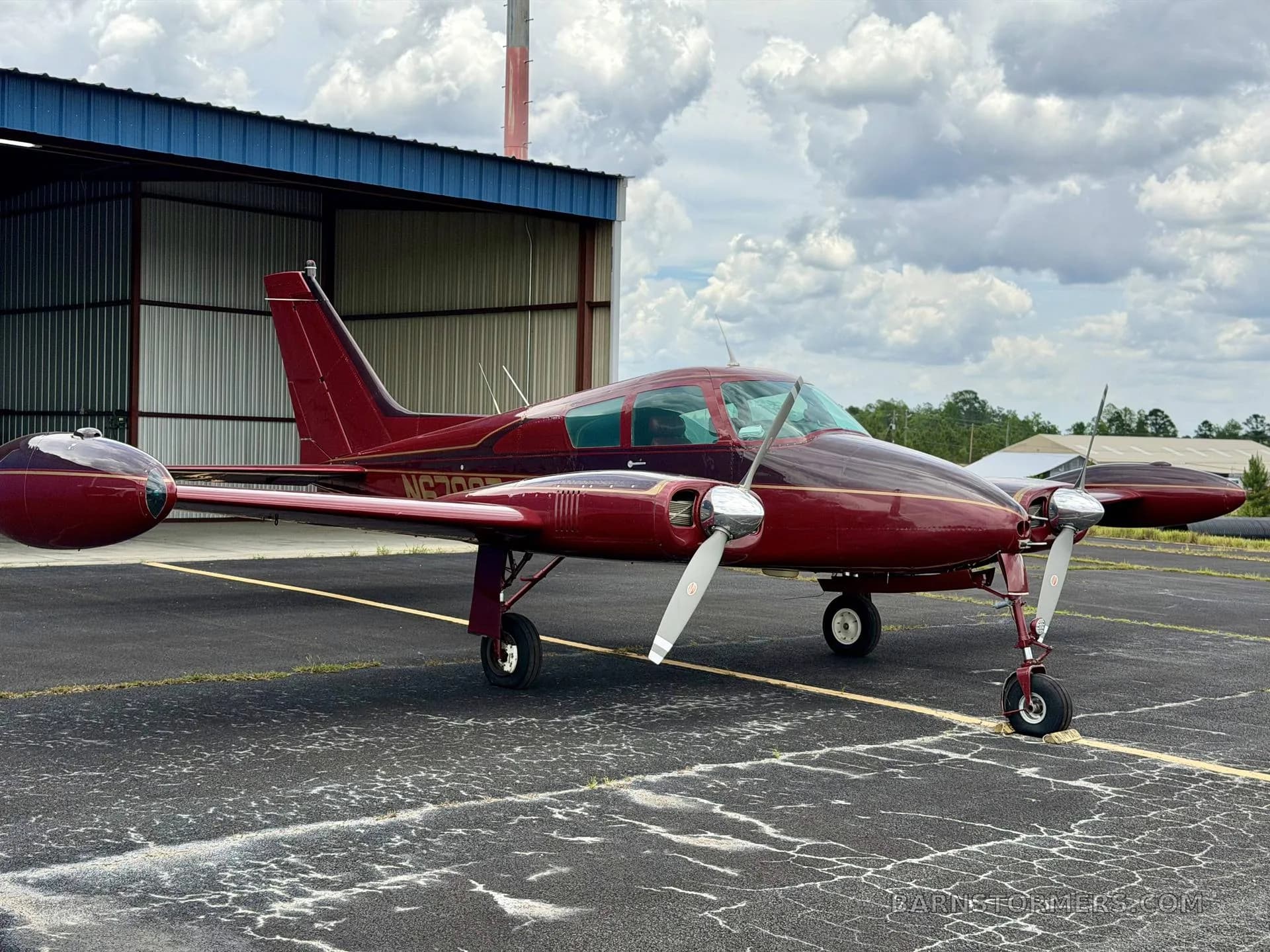 A red twin-engine aircraft, likely a Beechcraft Baron, with registration N6792T, parked on an asphalt apron in front of a blue hangar.