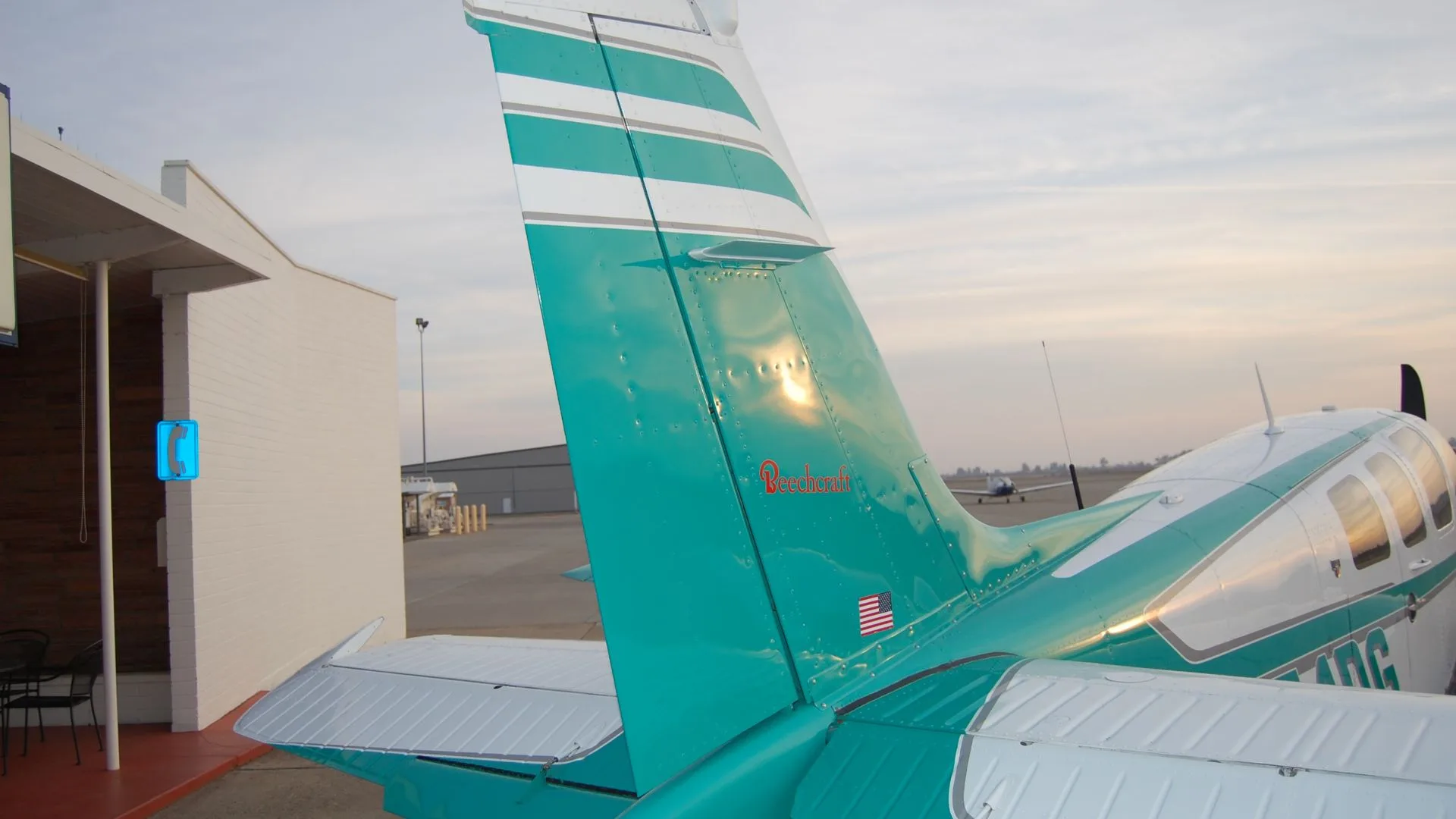The tail of the Beechcraft Bonanza N54DG, showing the teal and white paint scheme and the Beechcraft logo, with a building in the background.