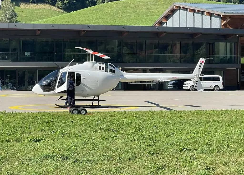 A white Bell 505 helicopter is parked on a tarmac with a person standing near its open door, against a backdrop of a modern building and green hills.