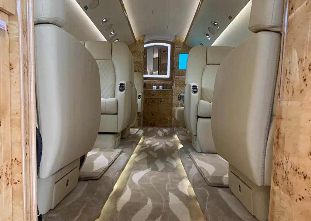 A view down the aisle of a private jet cabin, showing cream leather seats and a patterned carpet with light wood paneling.