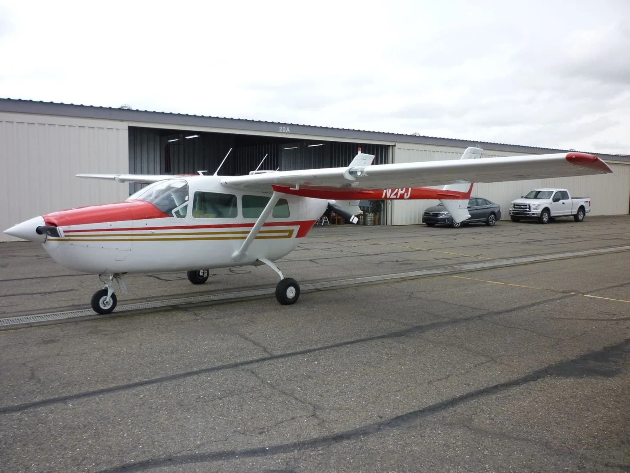 A front-side view of the white and red Cessna Skymaster N2PJ parked on an apron with cars and an open hangar in the background.