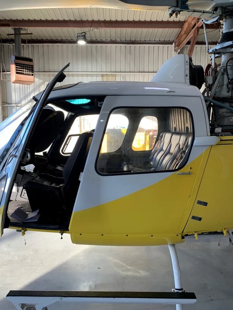 The side of a white and yellow Eurocopter AS350 B3 helicopter, N211FN, is shown inside a hangar with the pilot's door open.