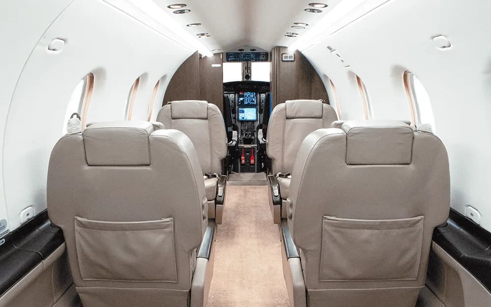 A view down the aisle of a private aircraft cabin, showing multiple rows of beige leather seats leading to the cockpit.