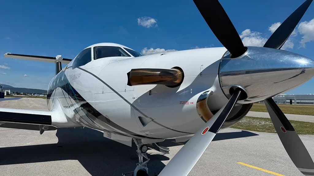 A close-up view of the front of a white Pilatus PC-12 aircraft, showing its propeller and engine cowling on an asphalt tarmac.