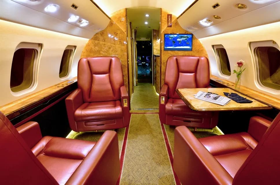 A wide view of the aircraft cabin interior with red leather seats, burled wood paneling, and a world map on the TV screen.