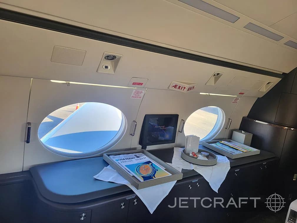 A close-up of a credenza inside the business jet cabin, featuring a monitor, magazines, and amenities.