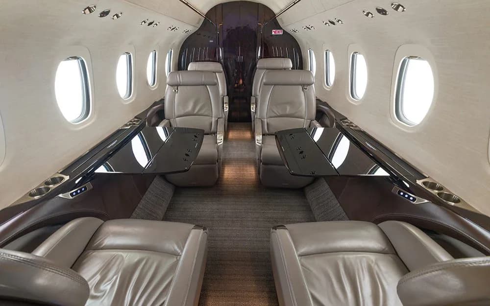 The interior cabin of a private jet, showing light brown leather seats with black fold-out tables deployed.
