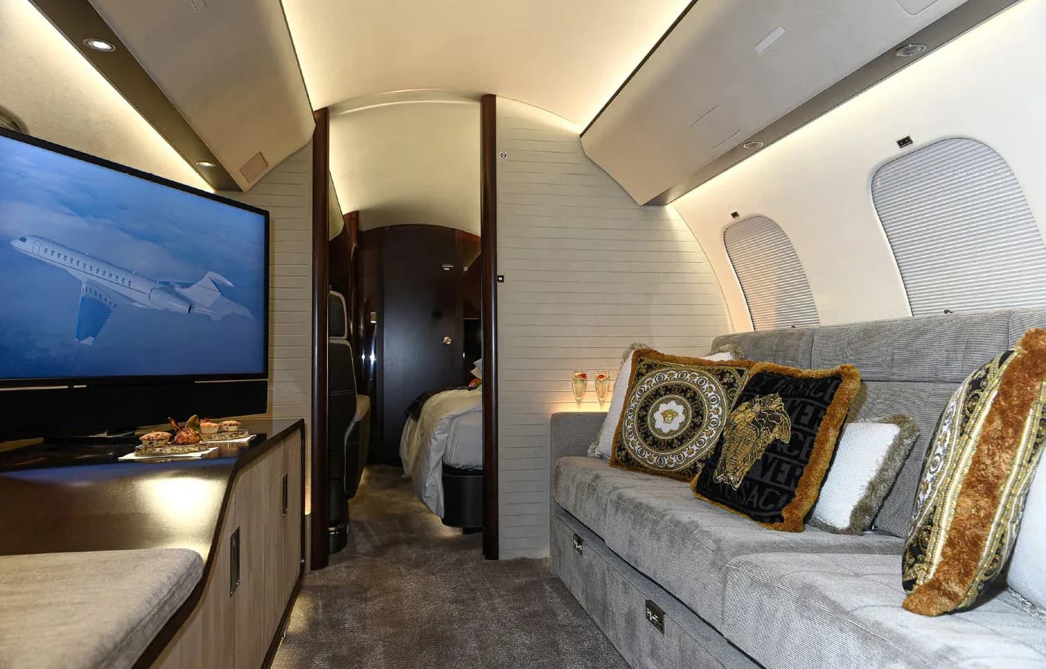 The private jet's lounge area with a grey sofa, Versace pillows, a large TV, and a view into the bedroom compartment.
