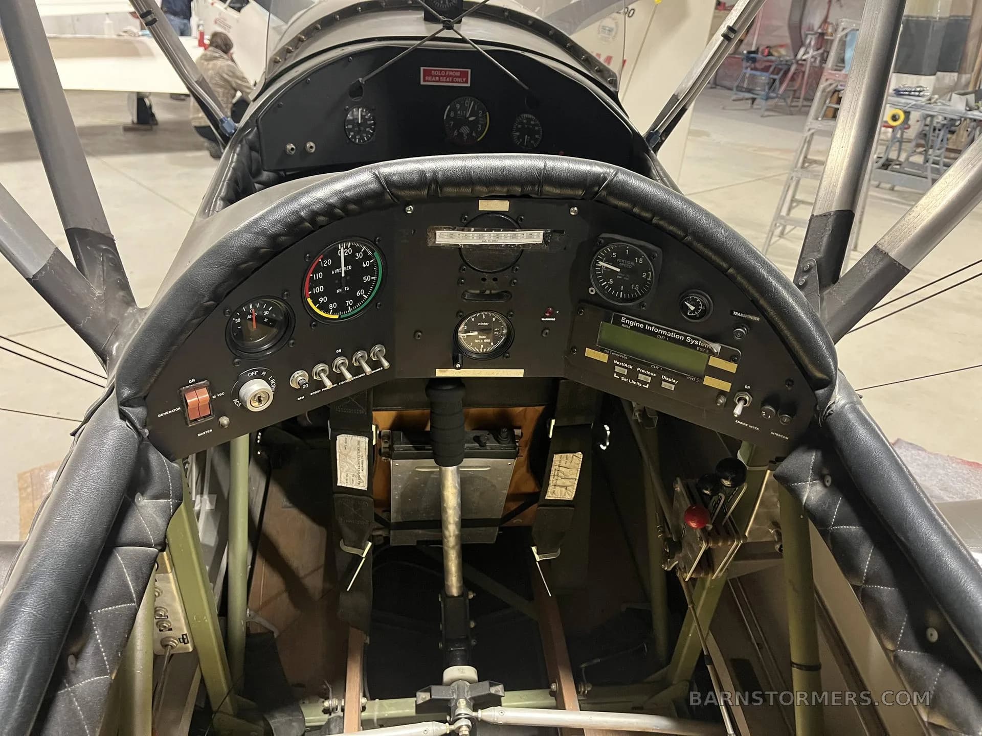 The cockpit of a biplane, showing the instrument panel with gauges, switches, and a control stick.