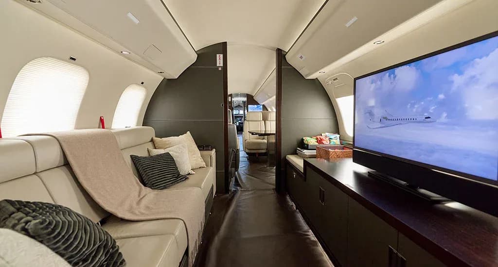 The aft cabin of a private jet with a cream leather sofa, throw pillows, and a large TV displaying an aircraft.