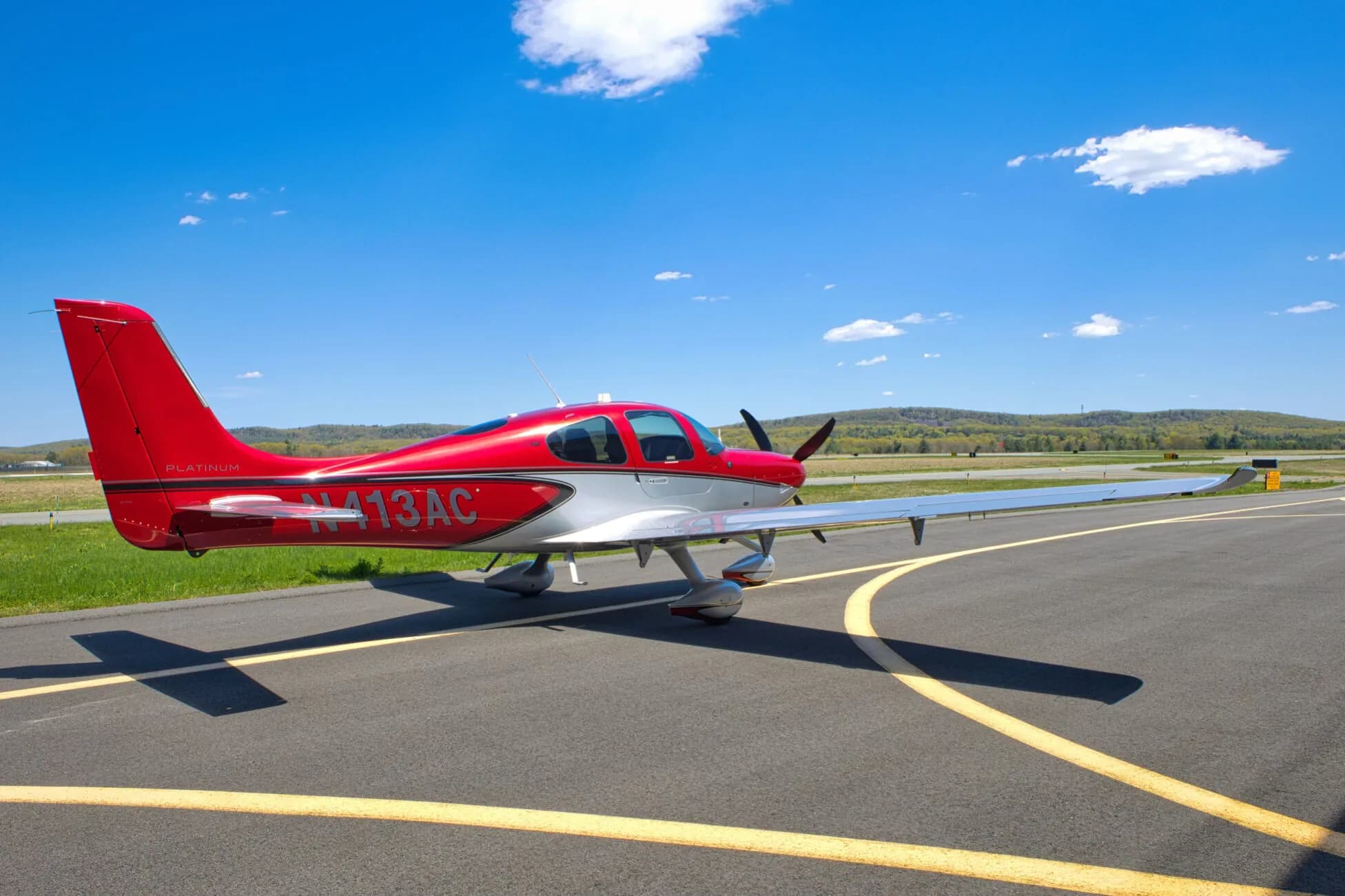 A red and silver Cirrus SR22T aircraft, N413AC, parked on an airport tarmac with hills in the background.
