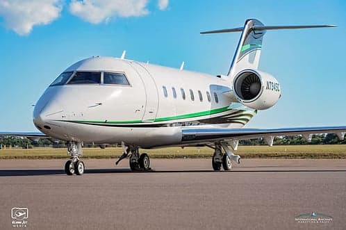 A white Bombardier Challenger business jet with green and black stripes, registration N794SC, parked on an airport tarmac under a blue sky.