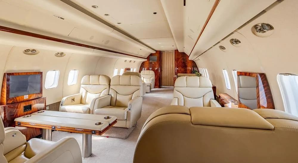 A luxurious aircraft interior with cream leather seats, wood tables, and polished wood paneling.