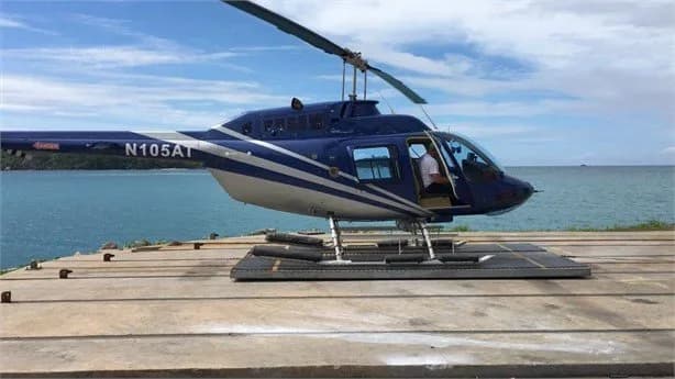 A blue and white helicopter, N105AT, is parked on a helipad near the ocean.