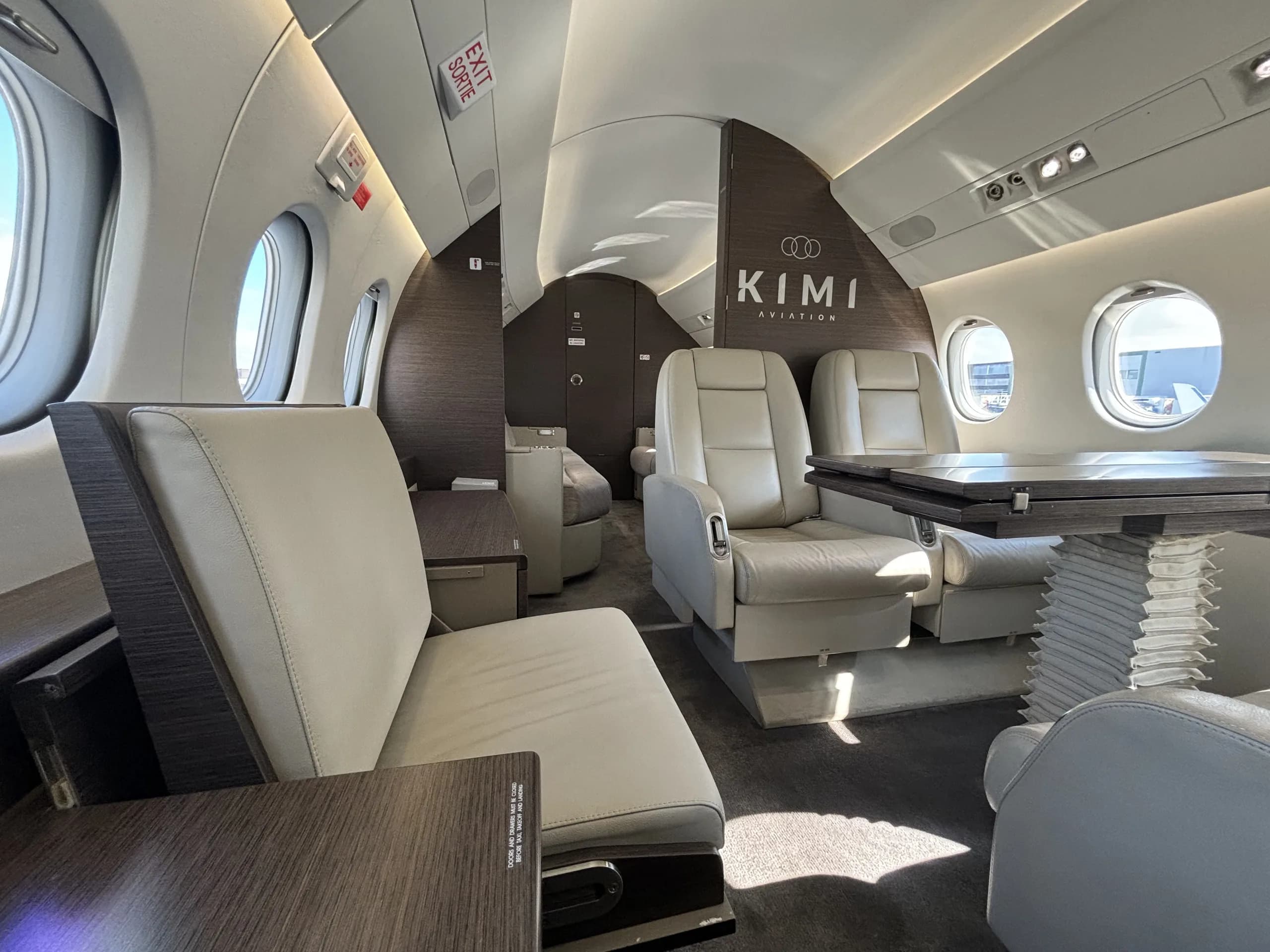 The luxurious interior of a private jet cabin, featuring light-colored leather seats and dark wood accents, with "KIMI AVIATION" logo visible.