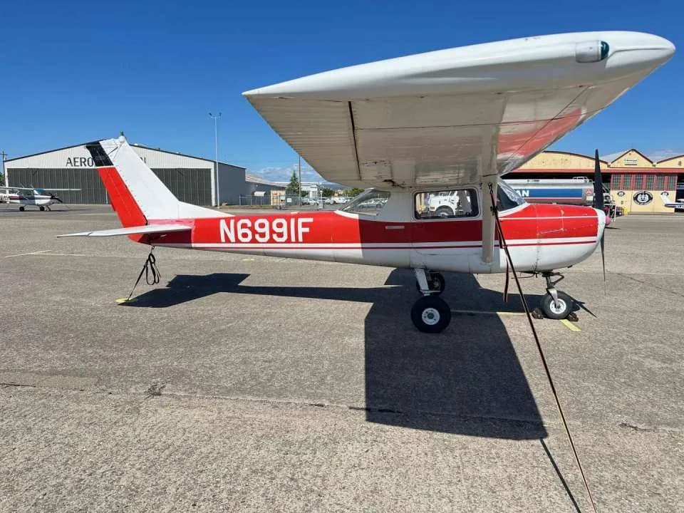 A red and white Cessna 150, tail number N6991F, parked on an asphalt apron with hangars in the background.