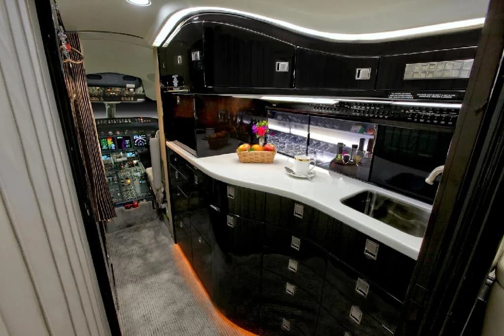 A well-appointed galley in the private jet, with dark glossy cabinets, white countertops, and a view into the cockpit beyond.