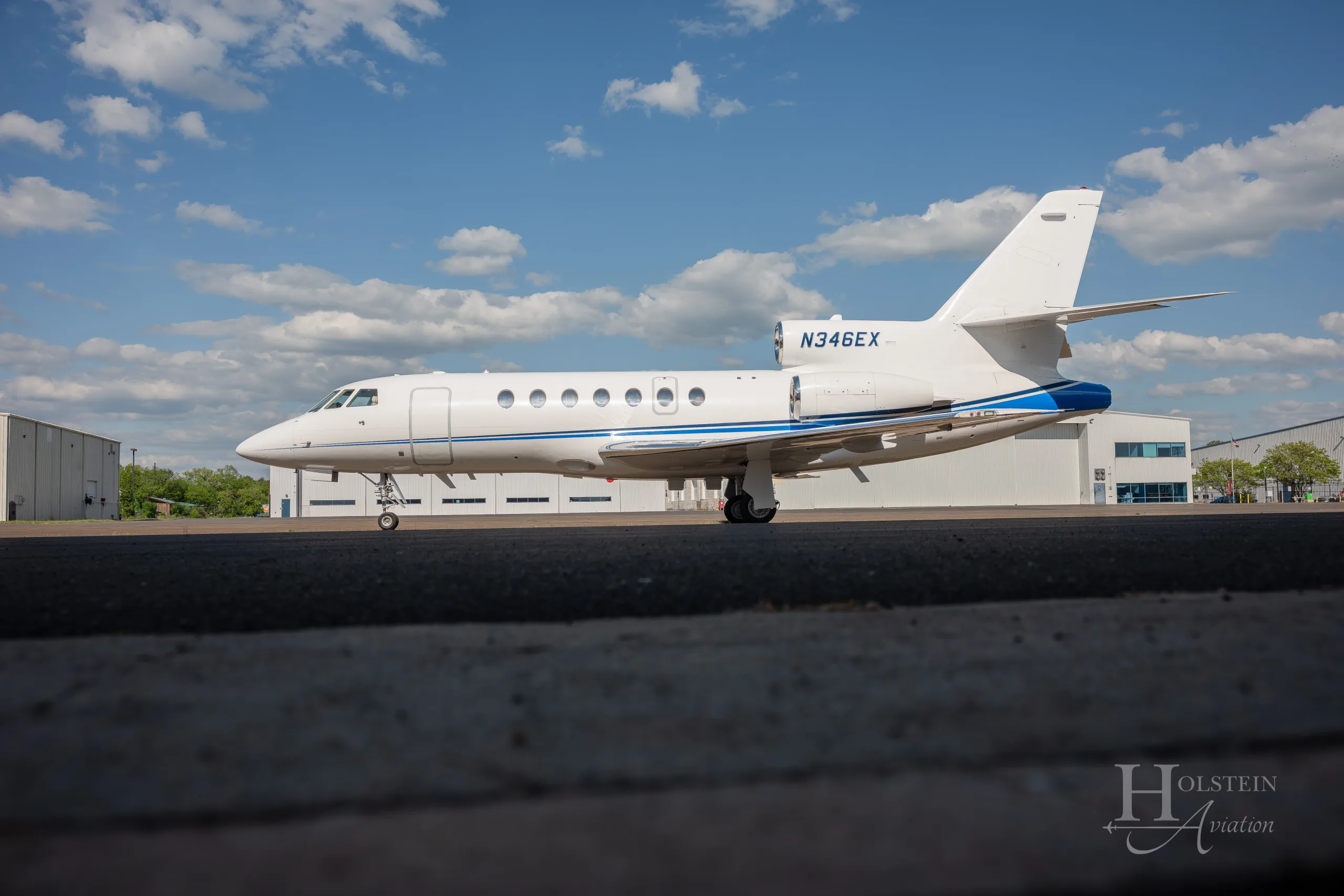 A side view of the white Dassault Falcon 900EX business jet, N346EX, with a blue stripe, parked on an airfield.