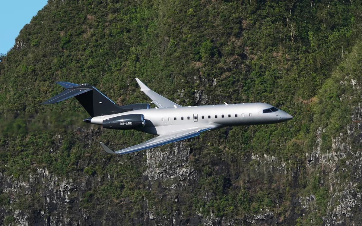 A silver and black Bombardier Global 6000 business jet with registration 9H-ARE flies past a lush green, rocky mountain.