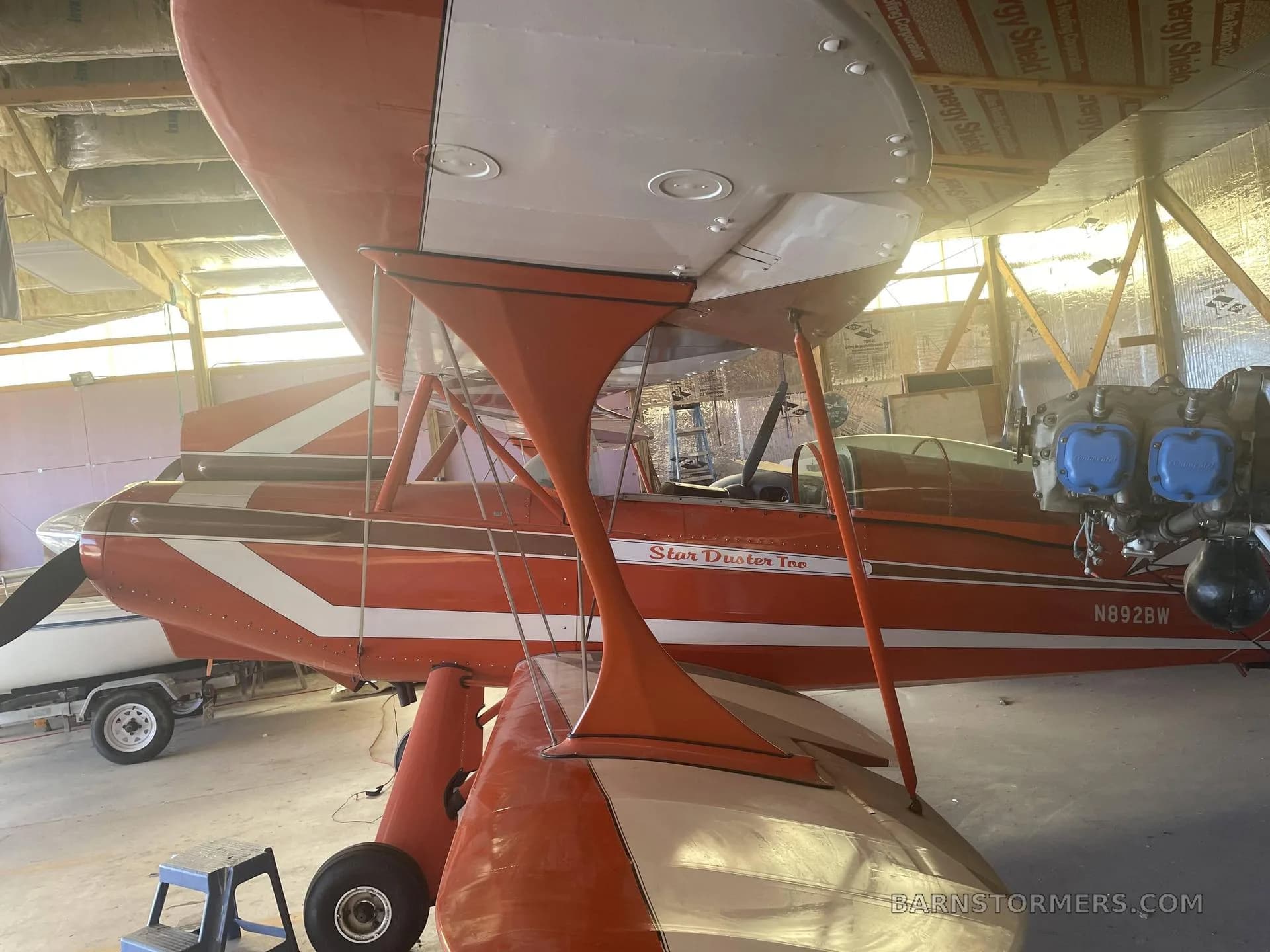 A side view of an orange and white Star Duster Too biplane with its engine exposed, parked in a hangar.