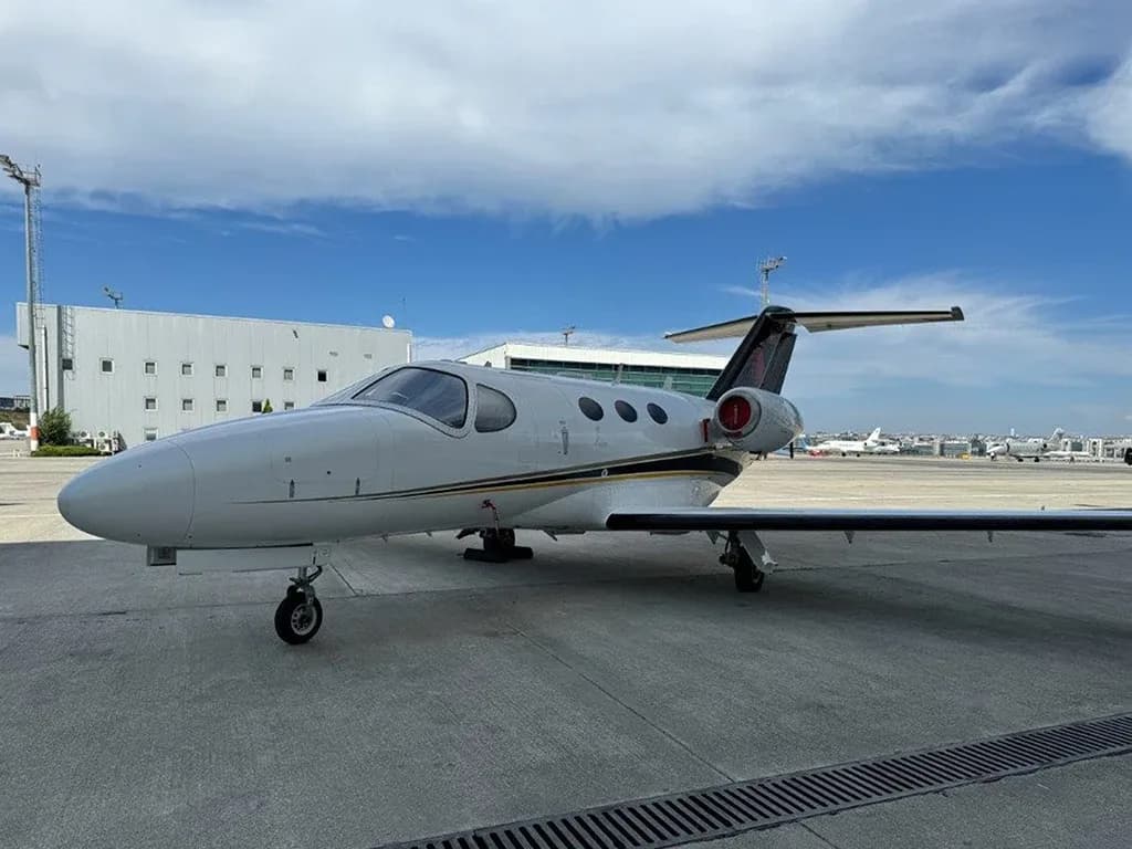 A white business jet with black and gold stripes on the tail, parked on an airport tarmac with buildings and other aircraft in the background.